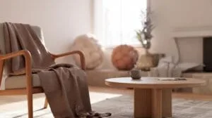 A light-filled living room features a mid-century modern style wooden armchair draped with a soft, brown throw blanket, positioned next to a round woo