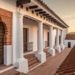 Colonial-style building with white walls, wooden doors, and a row of columns along a tiled walkway. Red clay roof tiles and distant mountains under a golden sunset sky are visible.