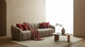 A beige channel-tufted sofa with red throw pillows and a matching throw blanket sits on a light rug in a minimalist living room.