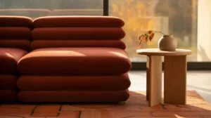 A plush, rust-colored sectional sofa with horizontal tufting sits on a terracotta-tiled floor next to a light wood side table holding a small, light-c