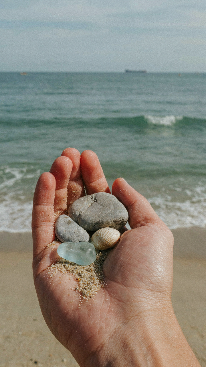 A hand holds beach treasures like sea glass and pebbles from a shoreline.