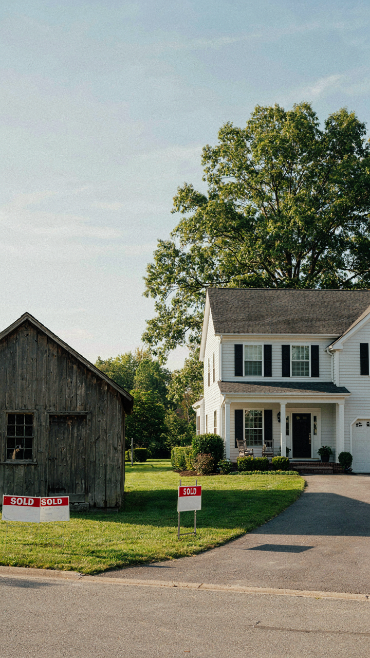 A 'Price Reduced' sign stands in front of a small barn house for sale.