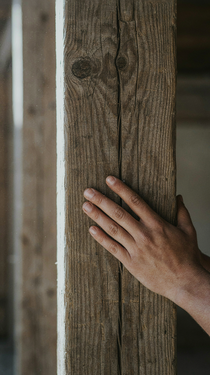 A hand touches the rough, textured surface of an old wooden beam, showing its grain.