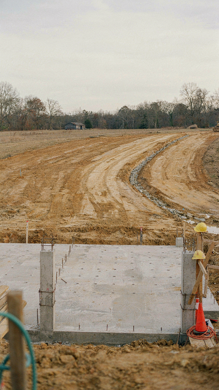 A construction site shows a farmhouse foundation with a porch and detailed drainage.