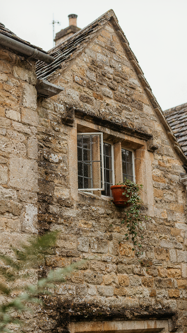 Rain-streaked honey stone Cotswold cottage wall with clustered windows.