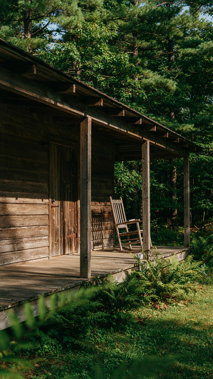 A wide wraparound porch creates deep shadows on cabin walls, protecting from sun.