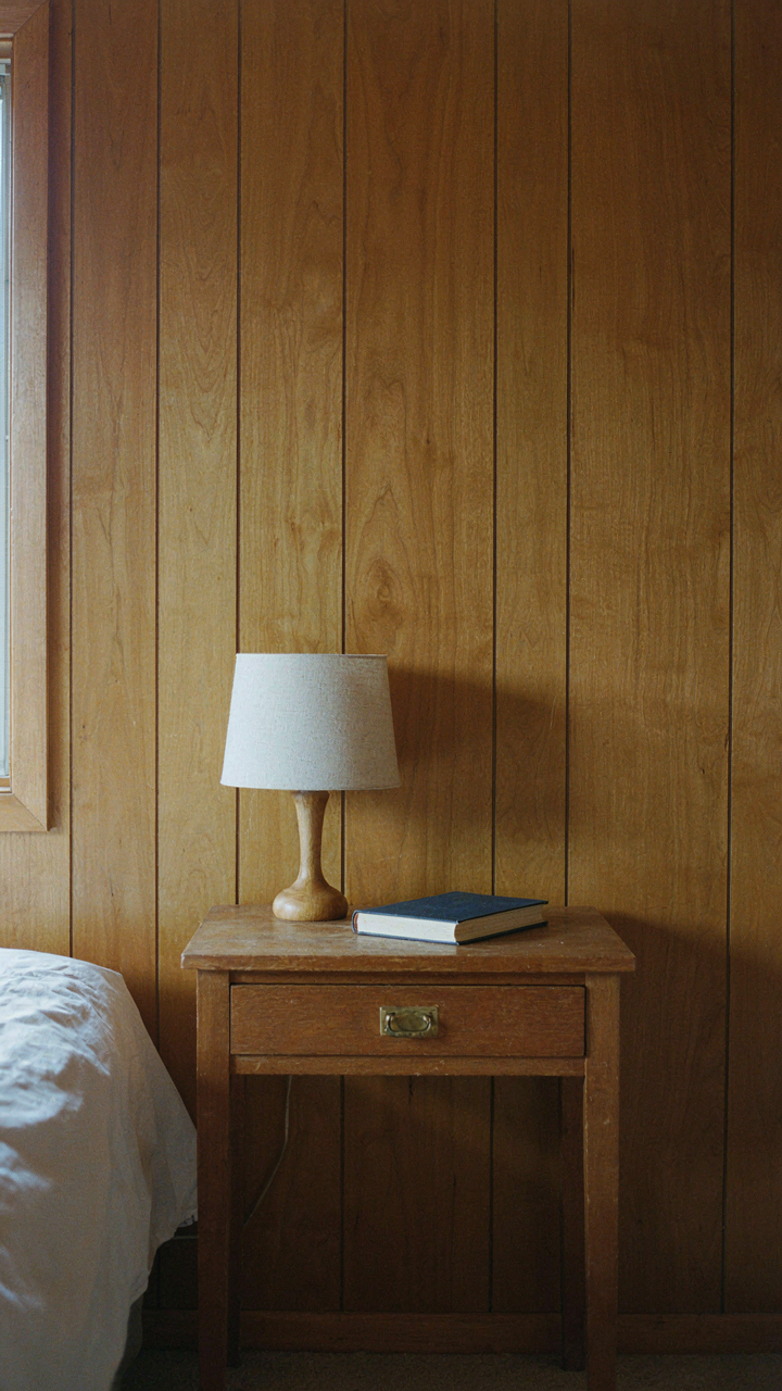 A simple nightstand holds a lamp and a book next to a clean wooden wall.