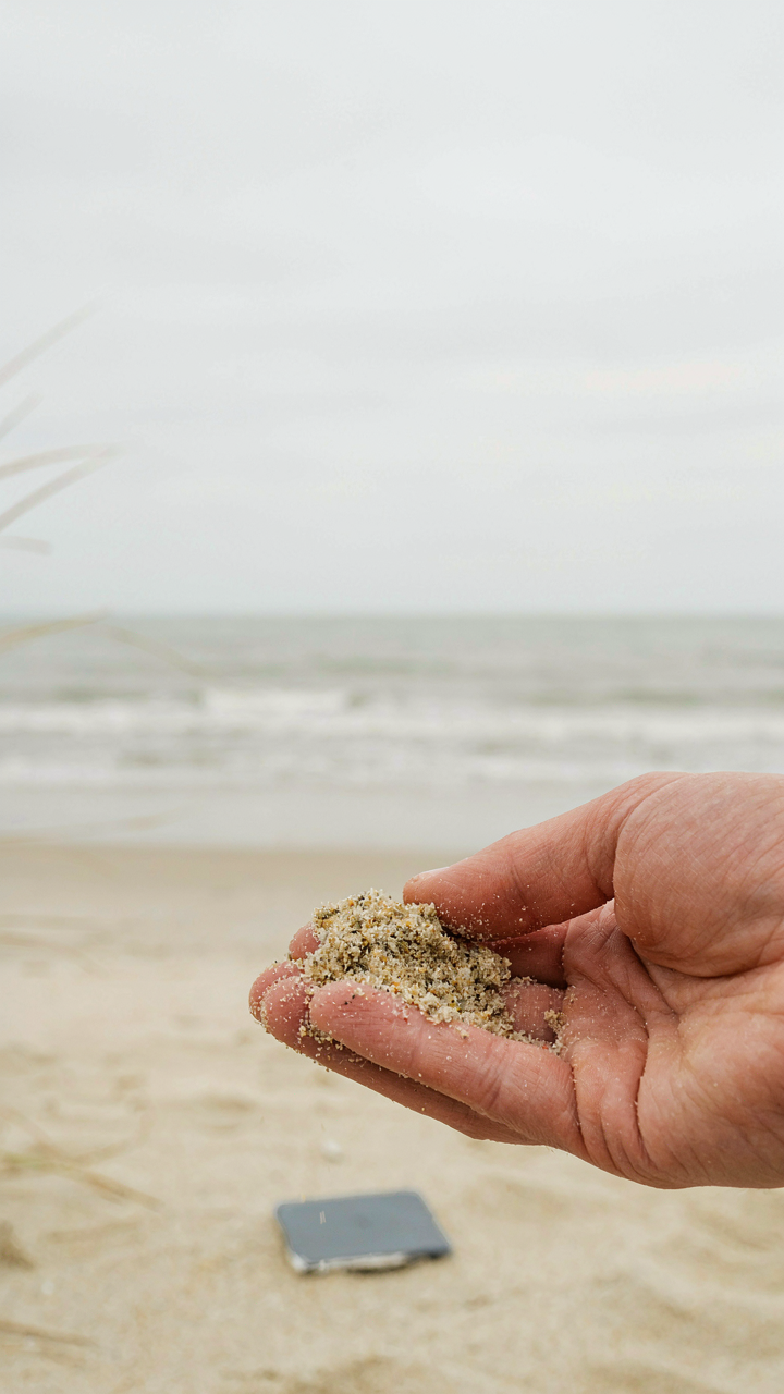 A hand holds sand next to a cool gray paint chip on a beach.