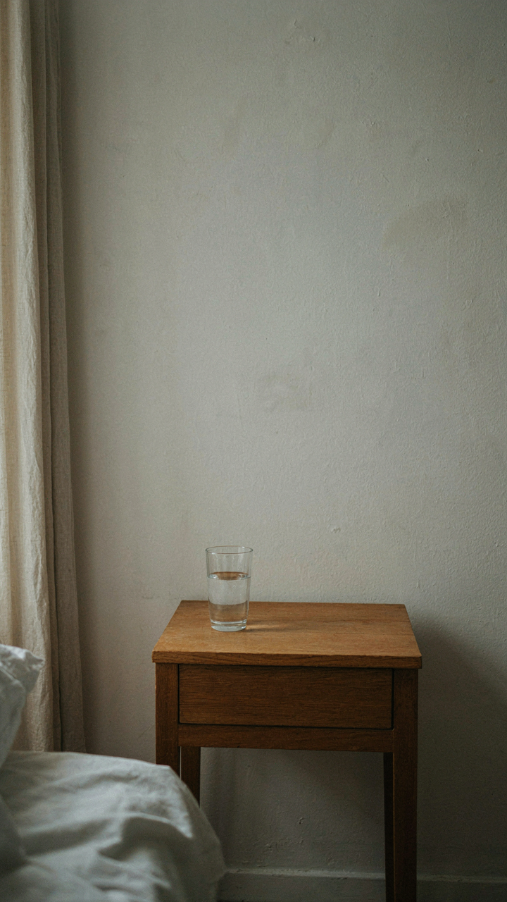 Minimalist wooden nightstand with a single water glass, empty wall, and cotton sheets.