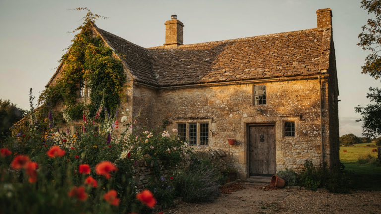 An old English farmhouse with stone walls and a green garden at sunset.
