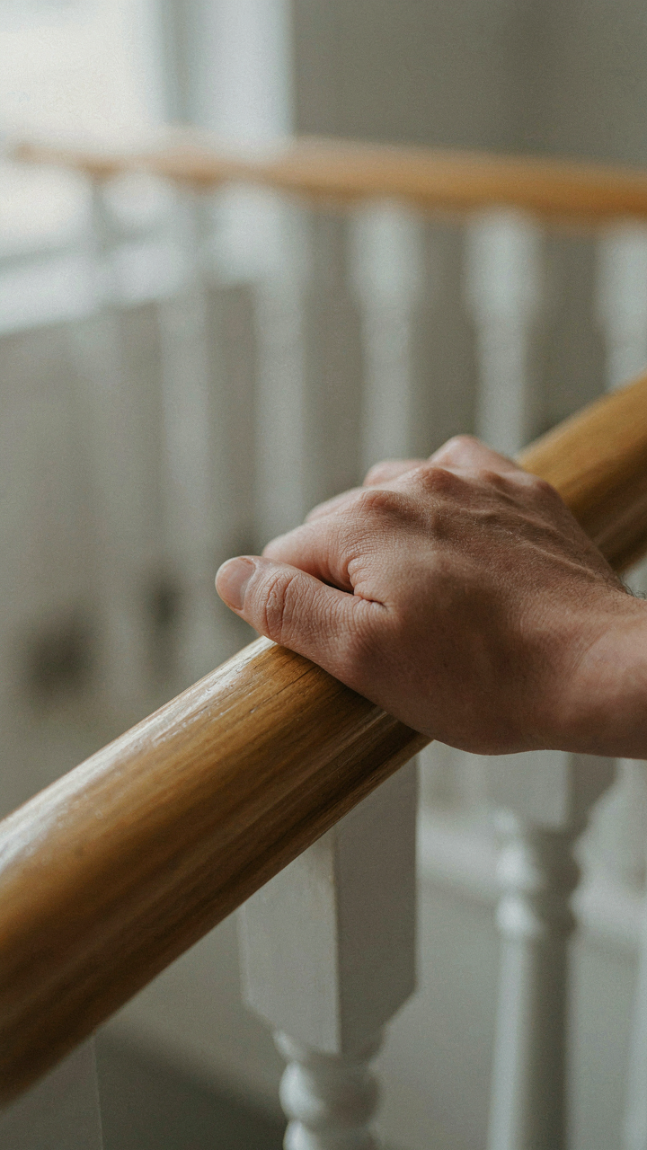 A hand grips a smooth wooden handrail on a farmhouse staircase.
