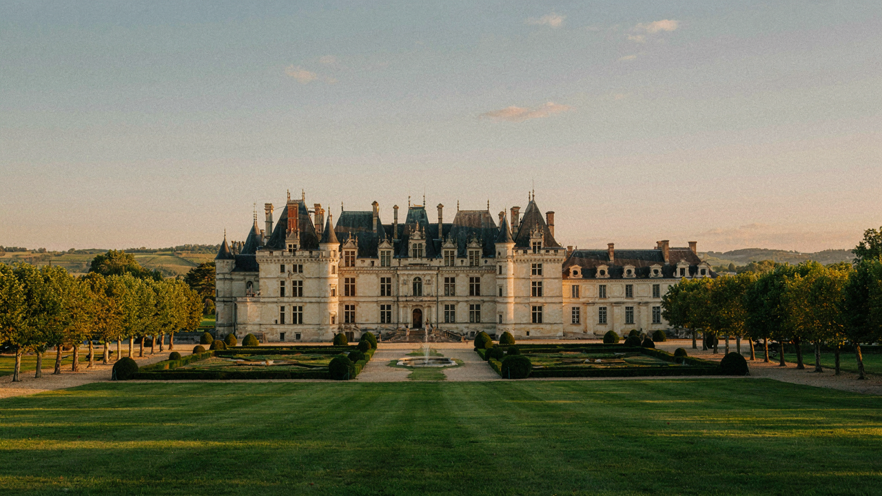 A grand French chateau with turrets stands under a golden sky, surrounded by manicured gardens.
