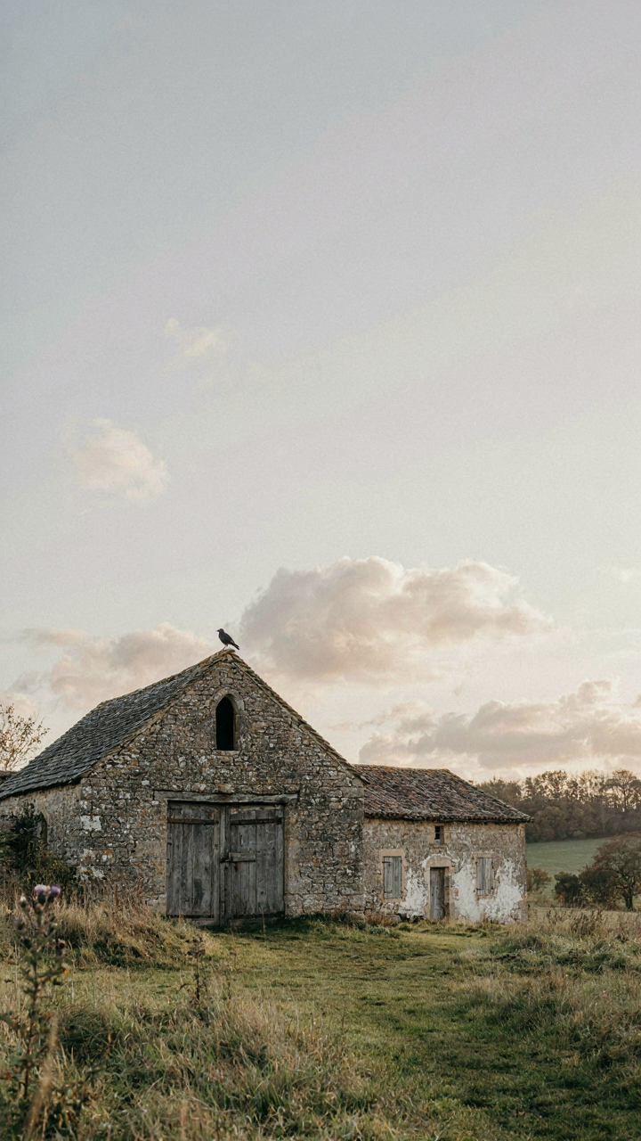 An old, weathered stone barn with timber doors stands next to a small stone farmhouse.