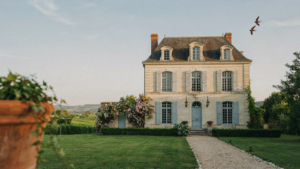 A beautiful french provincial house with a stone facade and a steep roof, surrounded by green trees.