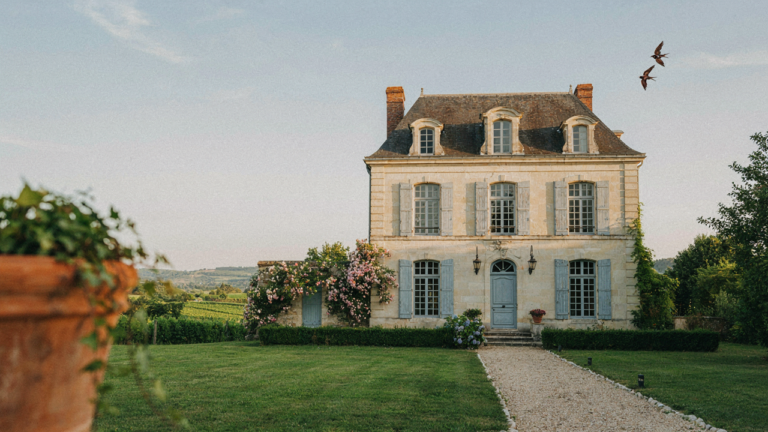 A beautiful french provincial house with a stone facade and a steep roof, surrounded by green trees.