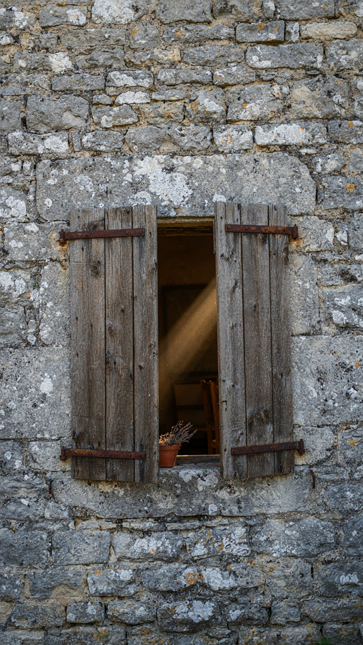 Sunlight streams through partially open weathered wooden shutters on a stone wall.