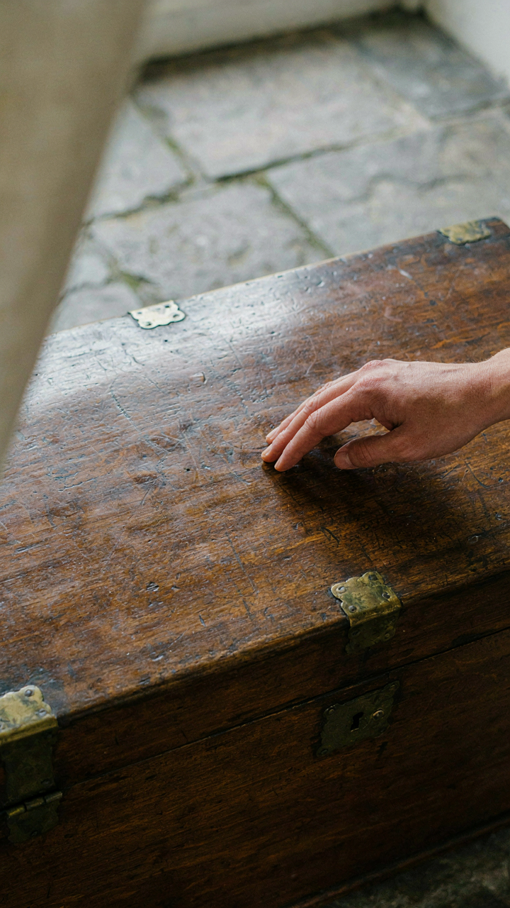 A hand touches the aged, dark wood of an antique chest on a flagstone floor.