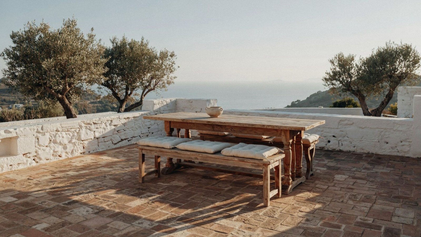 Sunlit coastal cottage terrace with an oak dining table and olive trees