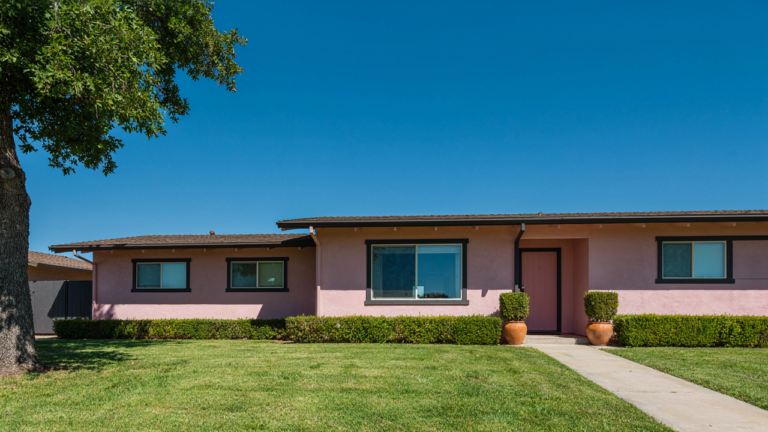 A soft pink mid century ranch home with dark contrasting trim under a clear blue sky.