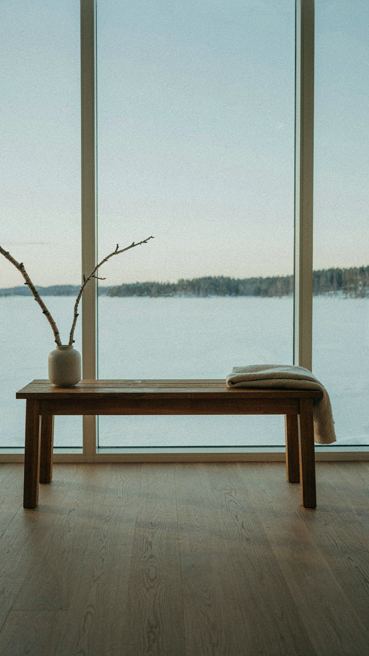 A lone wooden bench faces a large window with a view of a snowy lake.