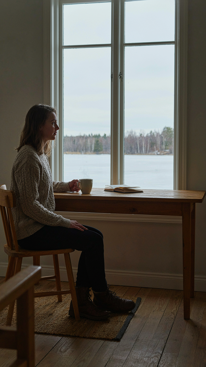 A person sits at a wooden desk, looking at a winter lake view from a large window.