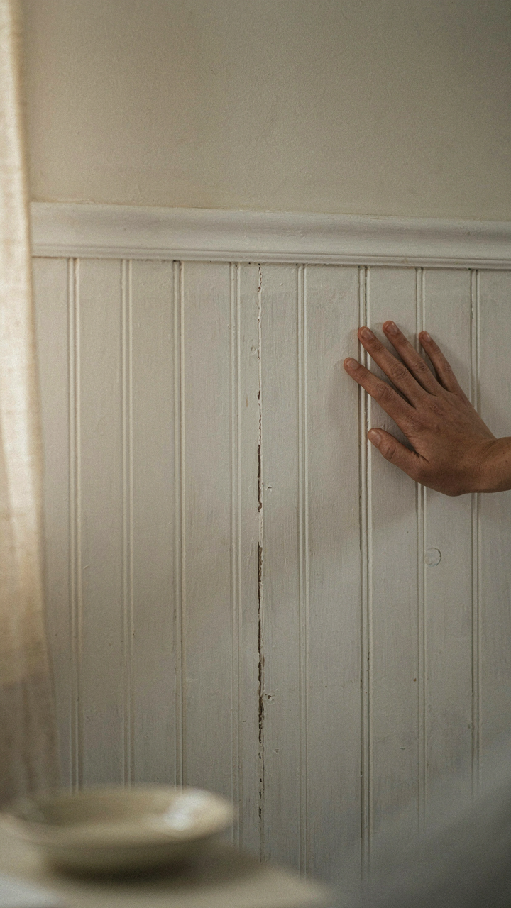 A hand touches a textured wainscoting wall in a cottage bedroom.