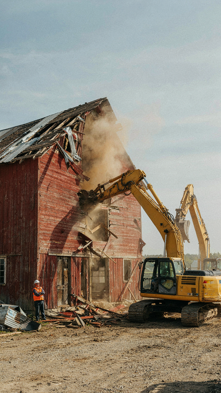 Heavy machinery demolishes an old barn, exposing its wooden beams.