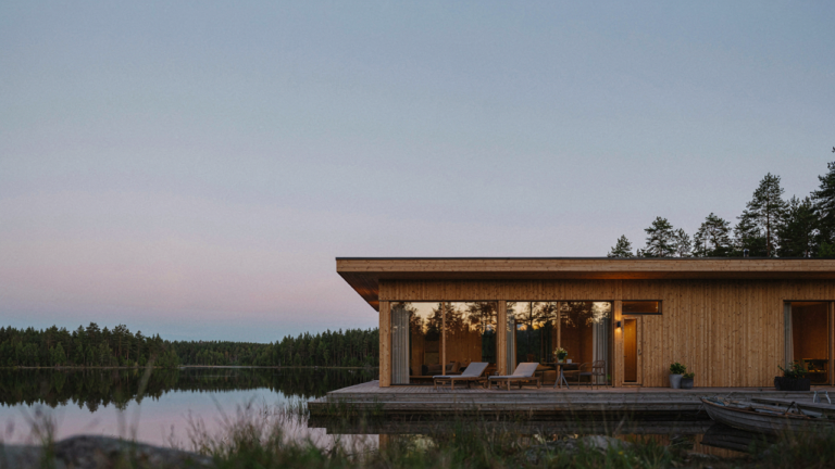 A modern Scandinavian lake house sits peacefully by a still lake at dusk, surrounded by pine trees.