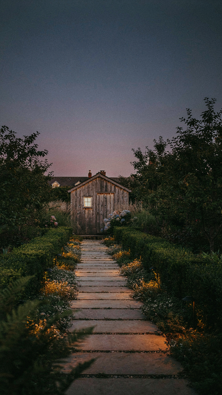 A softly lit garden path at dusk leads to a she shed, with low-level outdoor lighting.