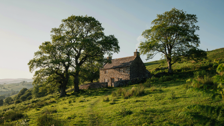 A rustic stone cottage sits nestled in a lush green hillside with smoke from its chimney.