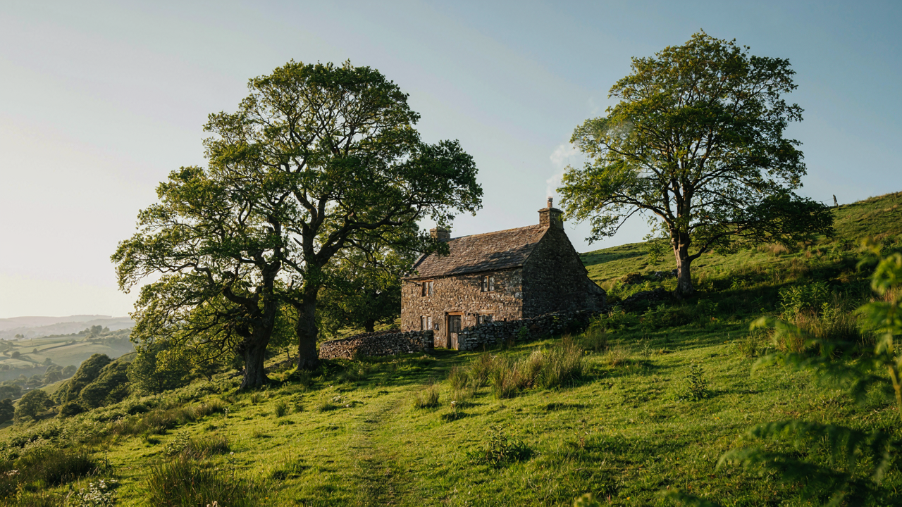 A rustic stone cottage sits nestled in a lush green hillside with smoke from its chimney.