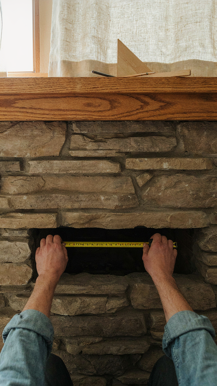 A hand uses a tape measure to check the width of a craftsman fireplace opening.