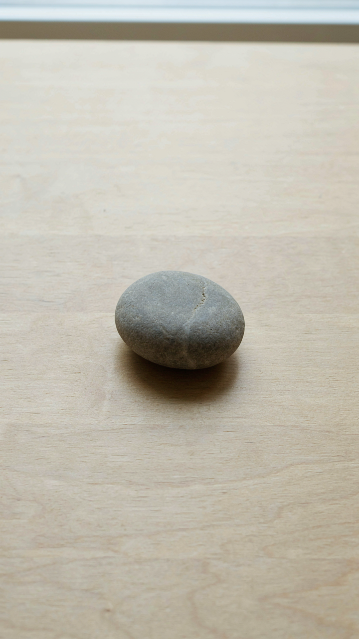 A single smooth grey stone rests on an empty light-colored wooden table.