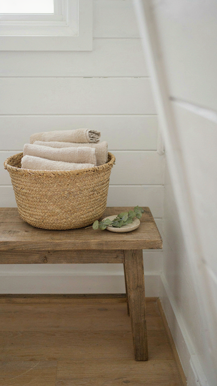 Rough wooden bench and seagrass basket with linen towels in a coastal bathroom.