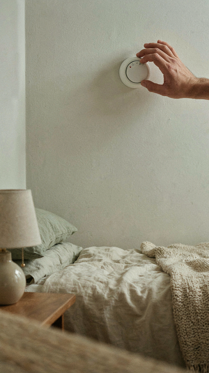 A hand adjusts a thermostat mounted on the wall next to a cottage guest bedroom bed.