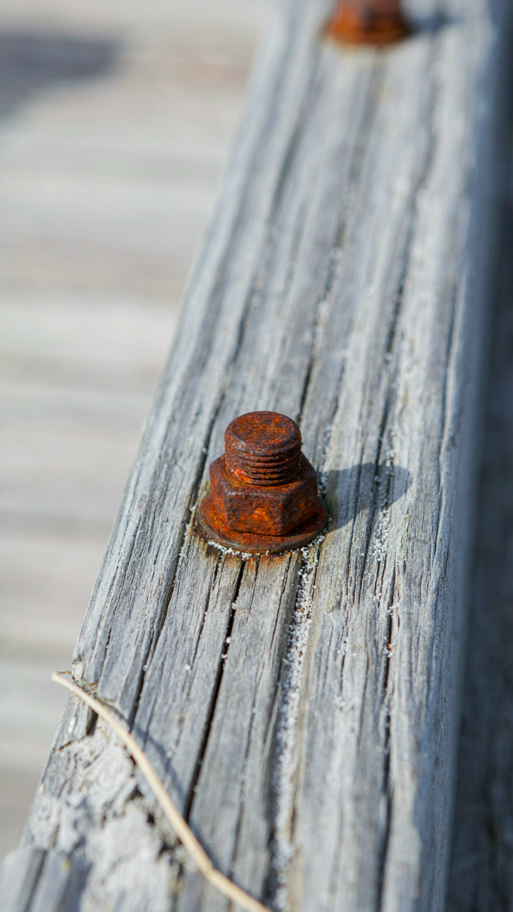 A rusted steel bolt connects wooden beams on a weathered coastal cottage porch.