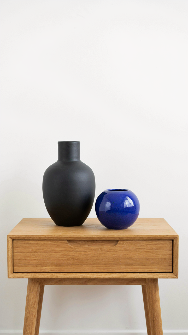 A single black ceramic vase on an empty light wooden nightstand.