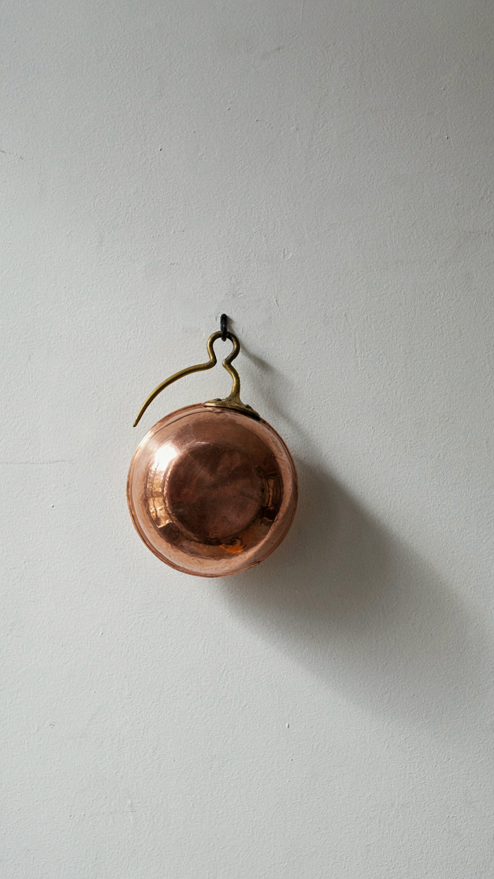 A single, shining copper pot hangs on a white wall.