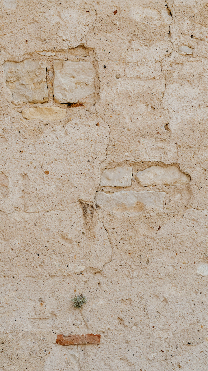 A close up of a weathered stucco wall with natural cracks and stone peeking through.