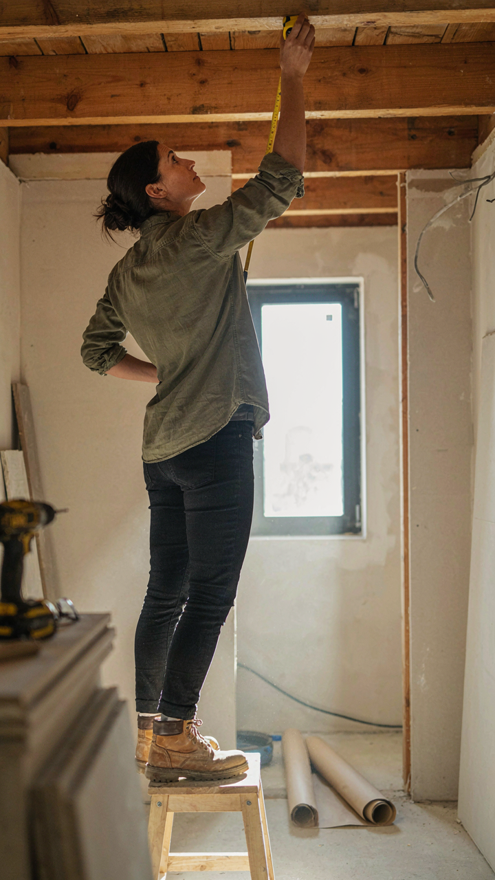 A person measures a low ceiling in a framed loft, considering space.