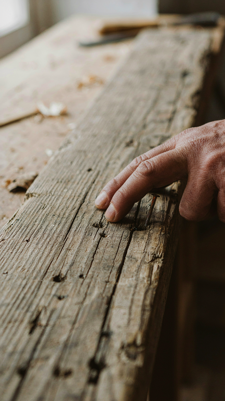 A hand touches a rough timber plank showing old nail holes and saw marks.