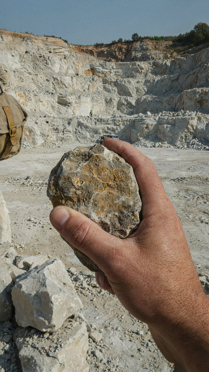 A person's hand holds a raw, local fieldstone against a quarry background.