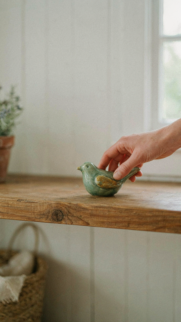 A hand places a ceramic bird figurine onto a rustic wooden farmhouse mantel.