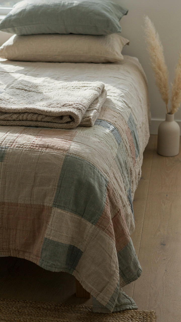 A bed with layered textiles, including a linen sheet, a wool throw, and a patchwork quilt.