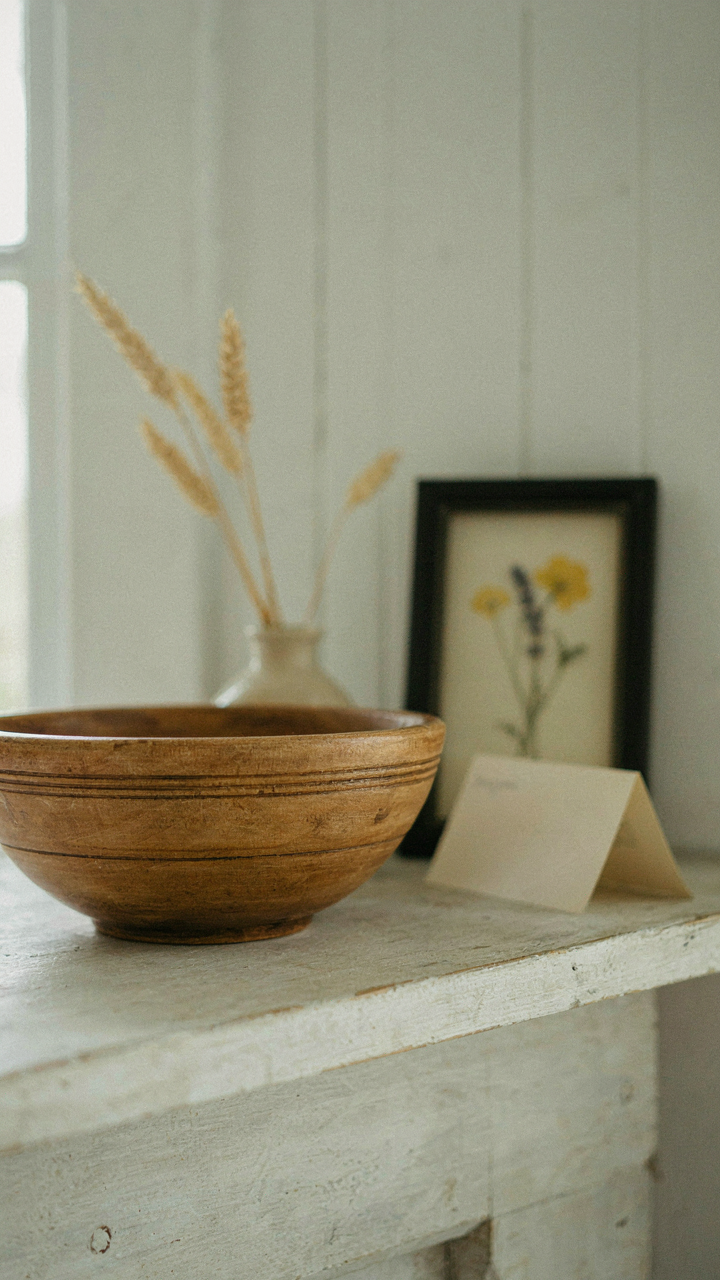 A vintage wooden bowl sits on a mantel next to a framed pressed flower and a recipe card.