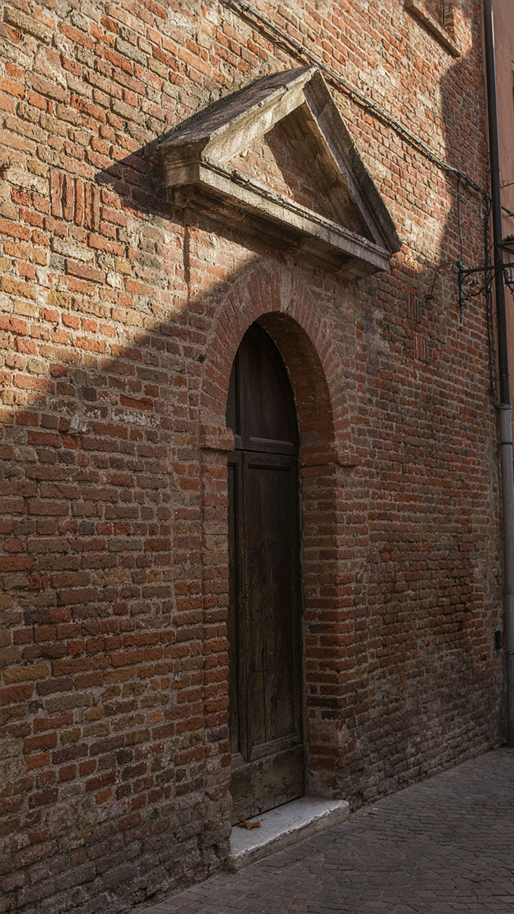 A heavy, slightly off-center pediment casts a strong shadow over a brick doorway.