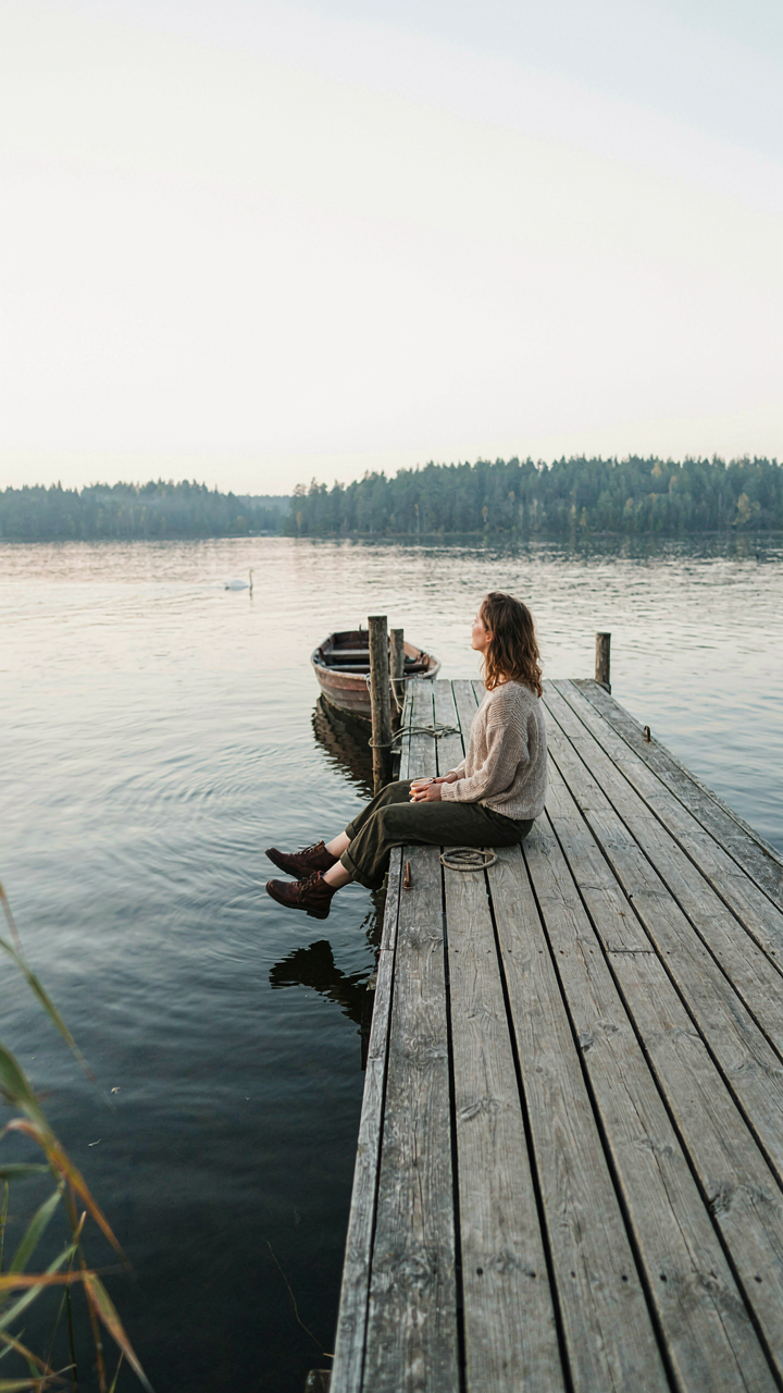 A person sits alone on a wooden dock looking out at a calm lake.