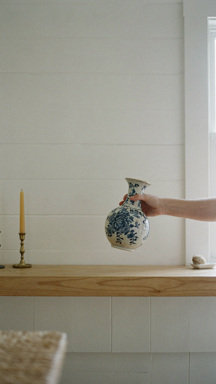 A hand removing an ornate vase from a simple wooden mantelpiece.