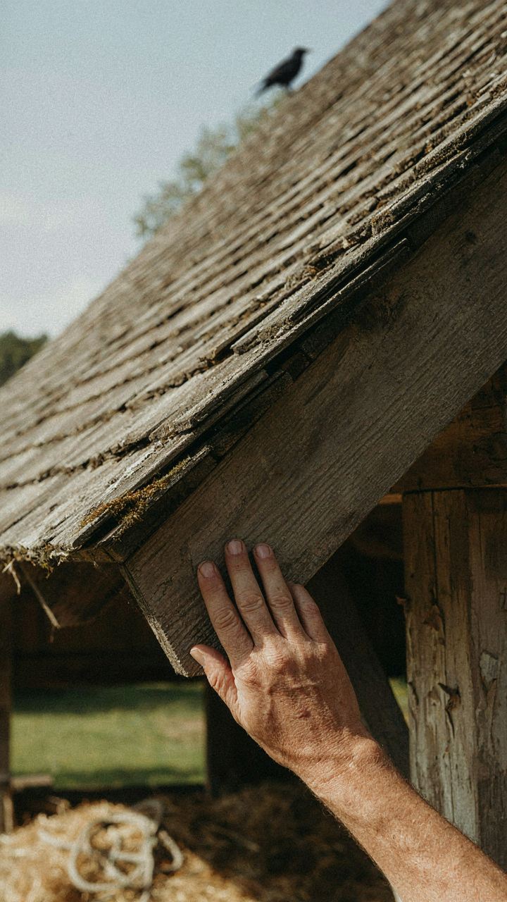 A person's hand touches the deep overhang of a steeply pitched roof on an old farm building.