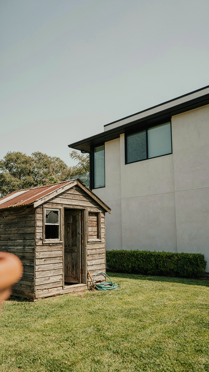 A small, dilapidated wooden shed next to a modern house.